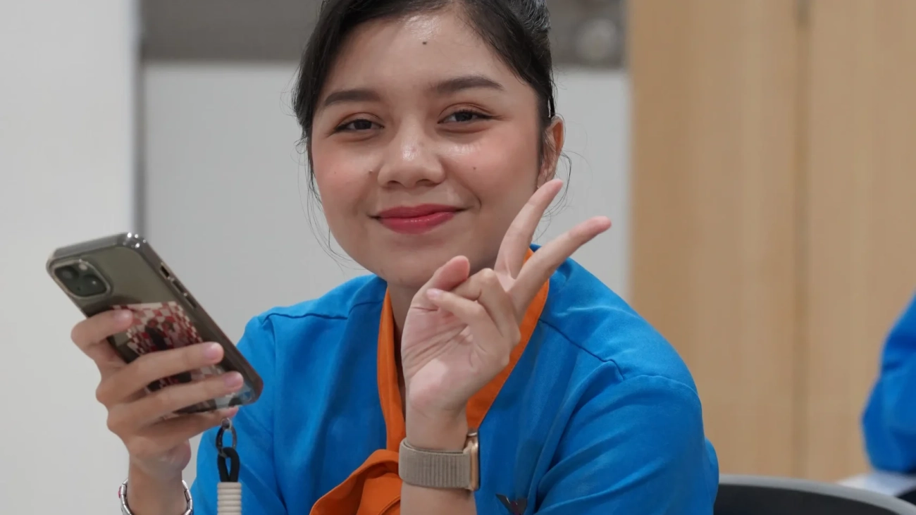 sekola penerbangan cepat lulus. Wanita muda berseragam biru oranye tersenyum, pegang HP, peace sign.