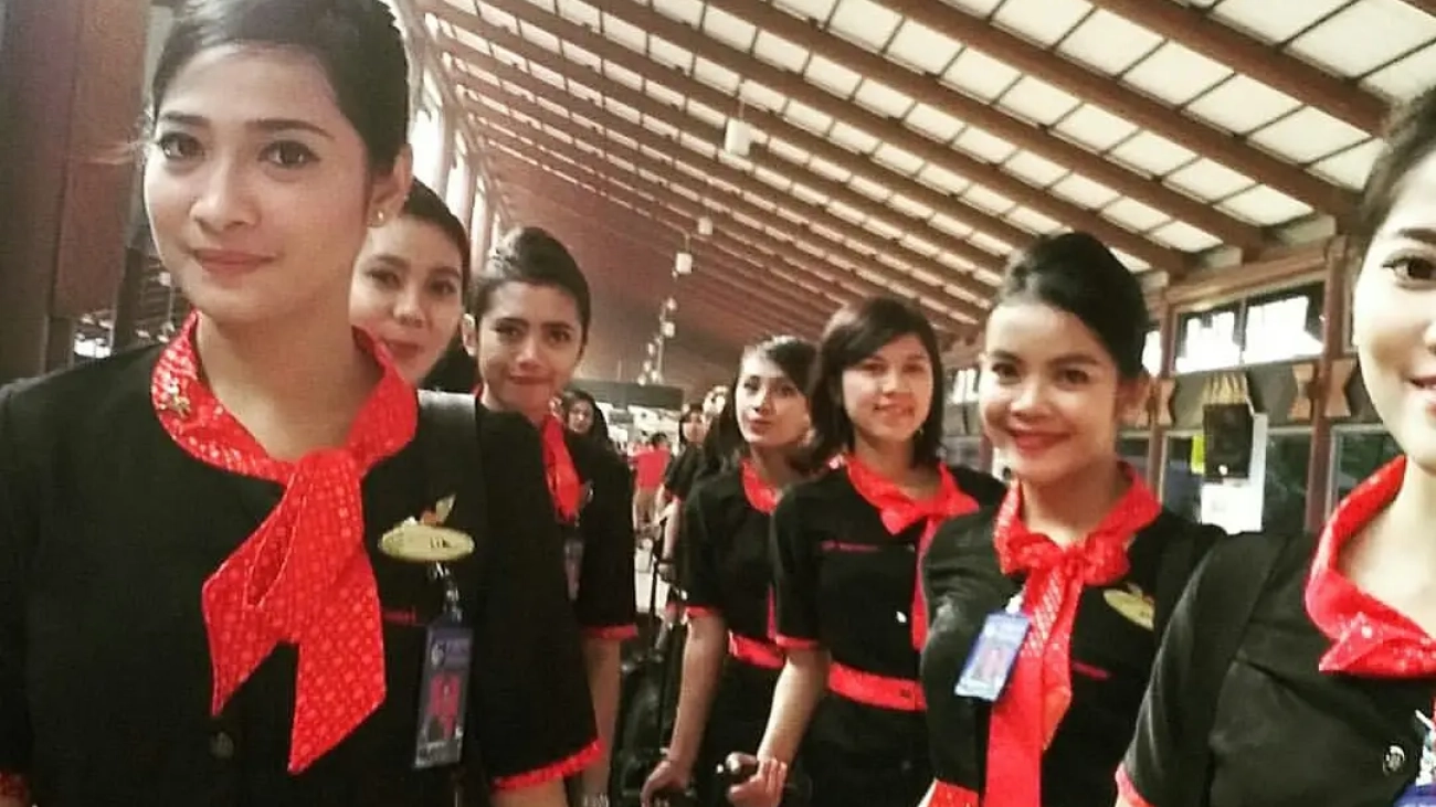 Cara Jadi Pramugari Profesional. Pramugari berseragam hitam merah tersenyum di terminal bandara.