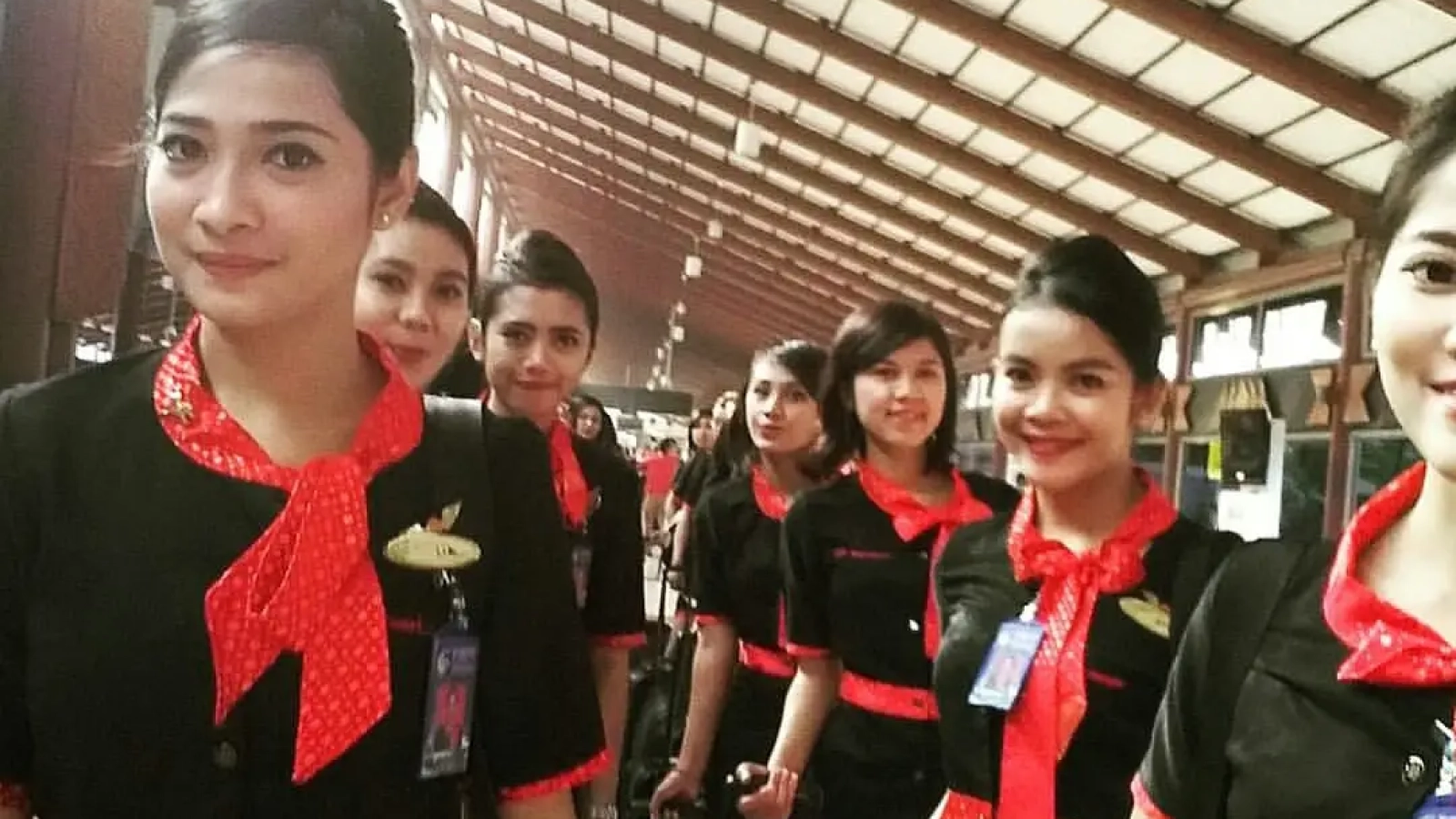 Cara Jadi Pramugari Profesional. Pramugari berseragam hitam merah tersenyum di terminal bandara.